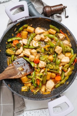 A vibrant stir fry featuring chicken, broccoli, snap peas, baby corn, and carrots dances in a sizzling skillet, with a trusty wooden spatula at the ready.