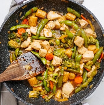 A vibrant stir fry featuring chicken, broccoli, snap peas, baby corn, and carrots dances in a sizzling skillet, with a trusty wooden spatula at the ready.