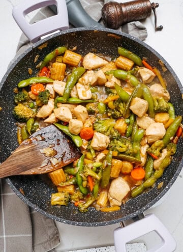 A vibrant stir fry featuring chicken, broccoli, snap peas, baby corn, and carrots dances in a sizzling skillet, with a trusty wooden spatula at the ready.