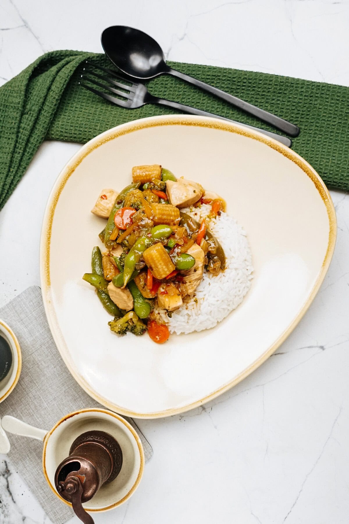 A tantalizing plate of stir-fry with vegetables and chicken rests elegantly on a marble surface, complemented by a serving of white rice. Beside it, a green cloth and spoon complete the inviting scene.