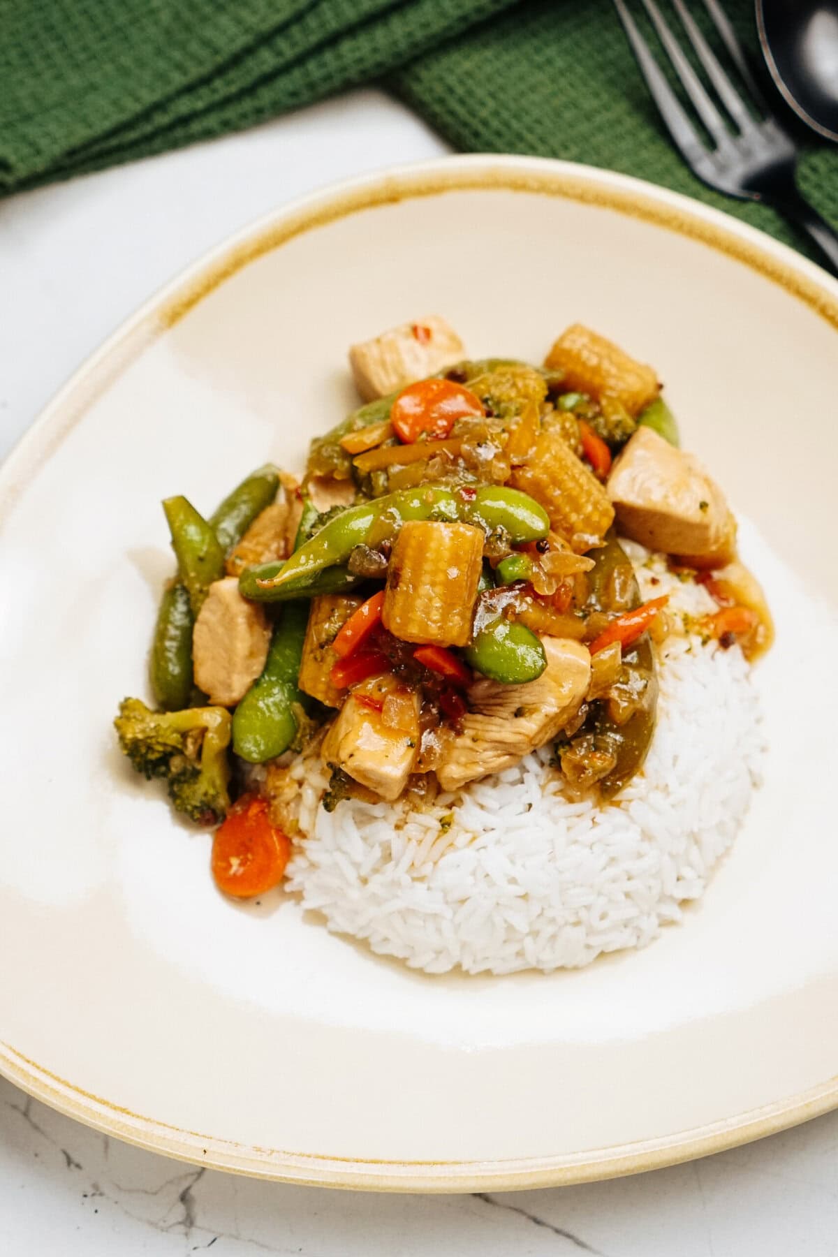 This vibrant stir fry dish features succulent chicken, crisp bell peppers, broccoli, snap peas, and baby corn served over a bed of white rice on an elegant cream-colored plate.