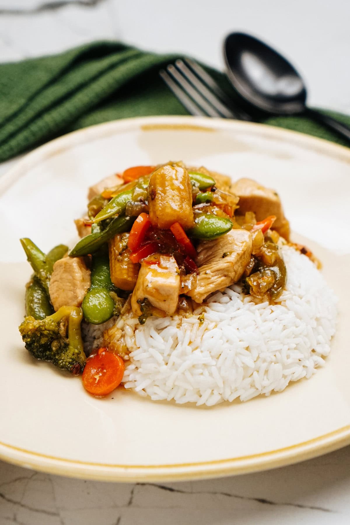 A plate of white rice topped with a savory stir fry of chicken, vibrant vegetables, and rich sauce. A fork and spoon rest on a green napkin in the background.
.