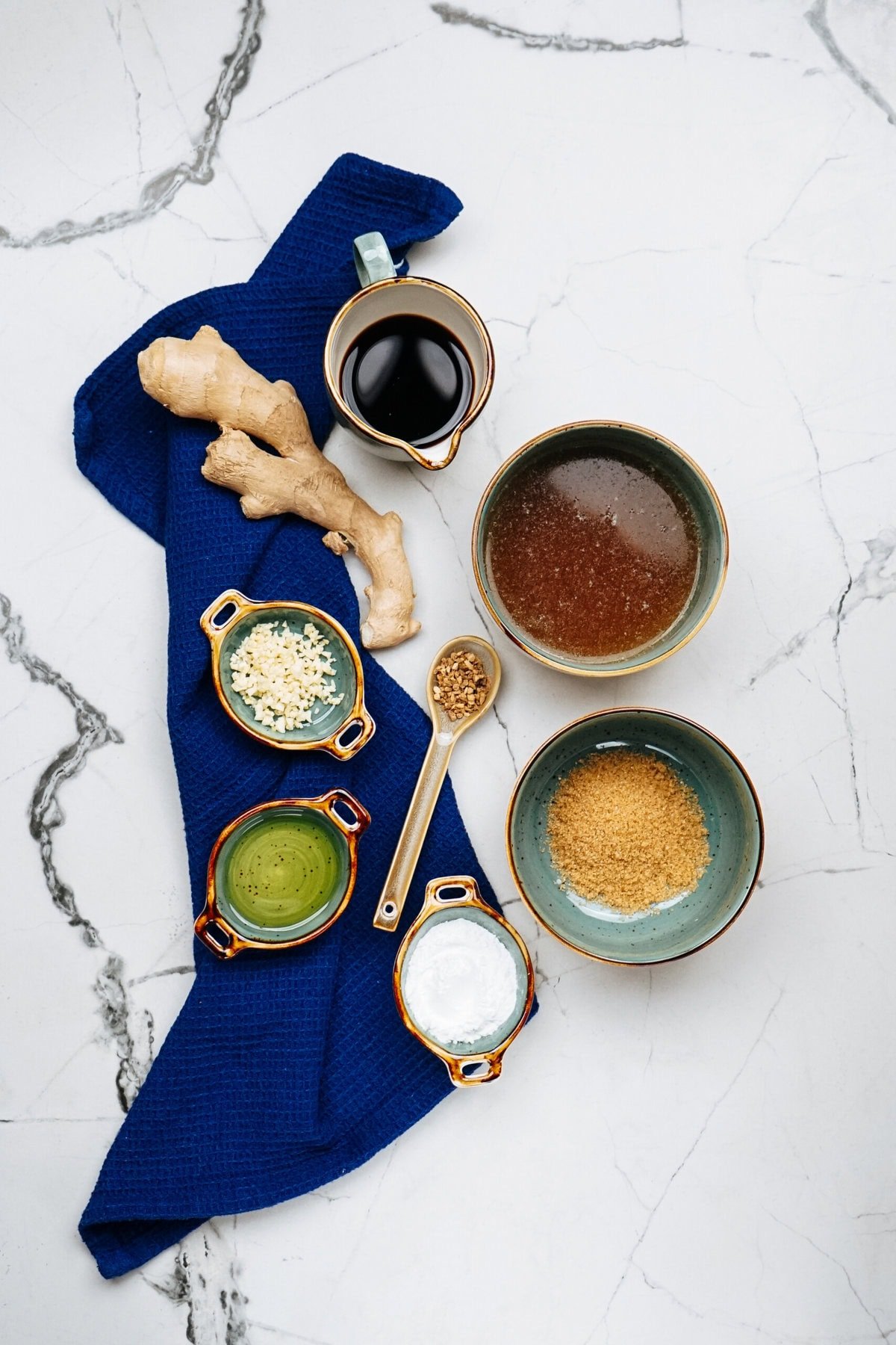 Ingredients arranged on a marble surface: ginger root, soy sauce, brown sugar, minced garlic, crushed pepper, oil, and salt—all set on a blue cloth with a wooden spoon. Perfect for enhancing your Sausage Alfredo with an unexpected twist of flavor.