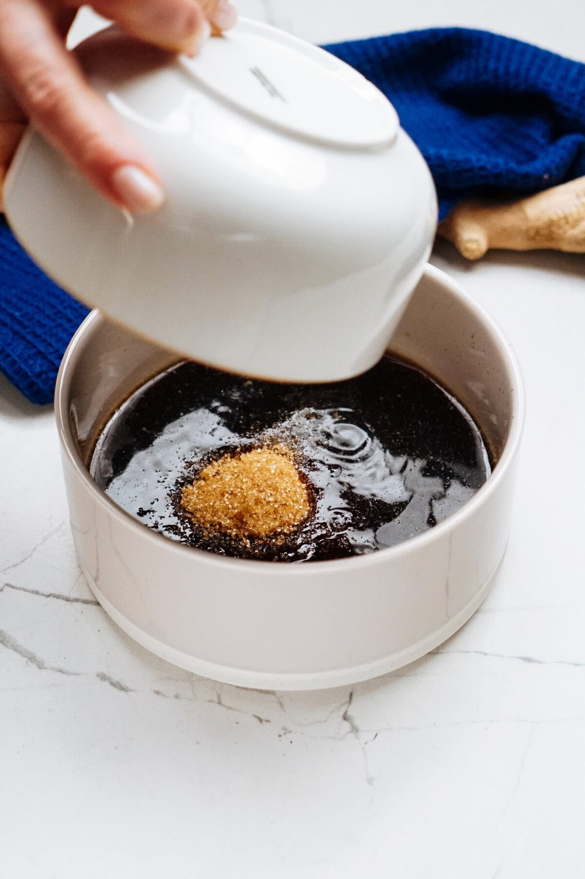 Brown sugar being added to a bowl of dark liquid on a white countertop, like preparing a sweet sauce for a stir fry, with a blue cloth in the background.