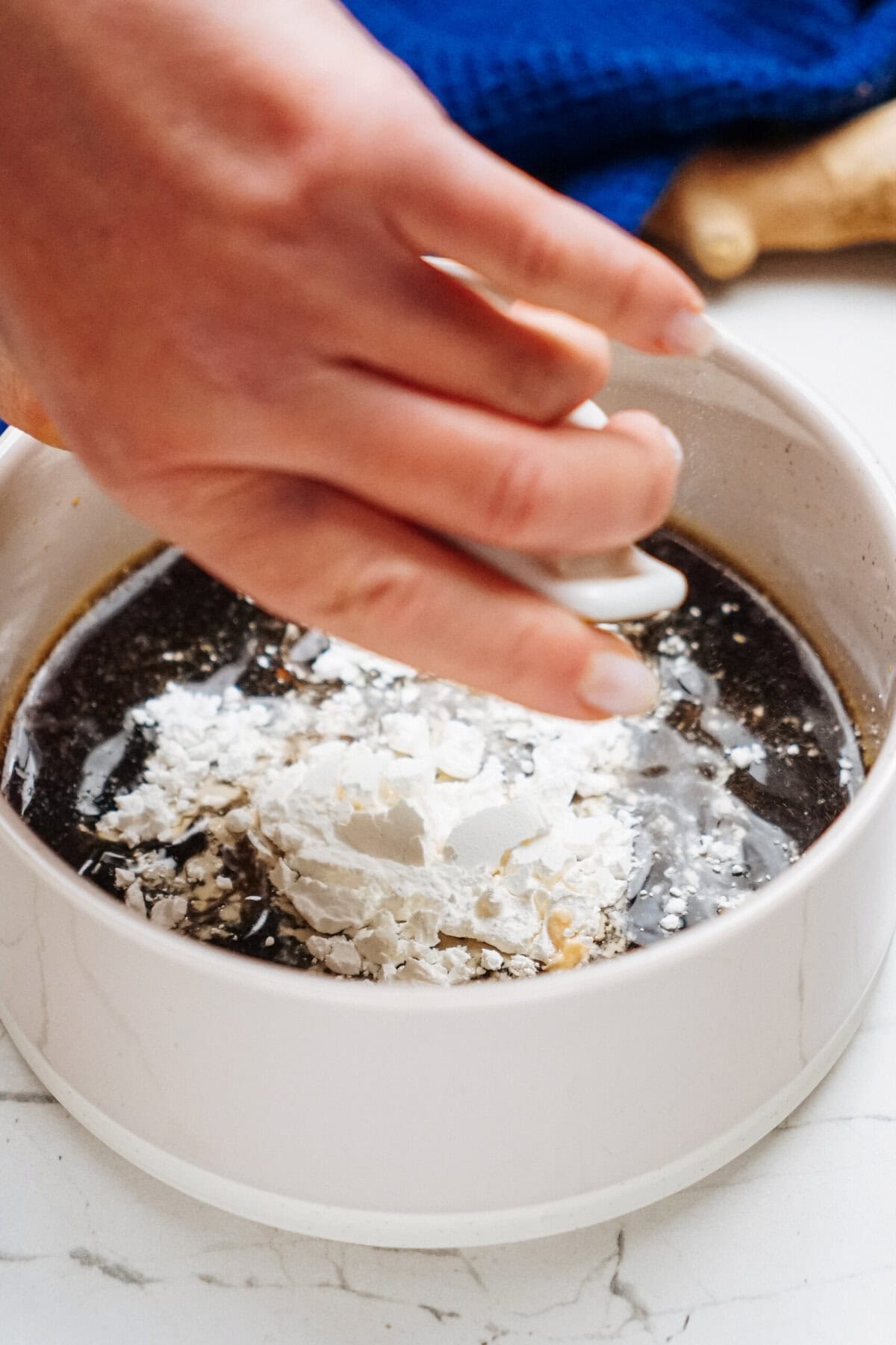 A hand gently adds white powder to a bowl of dark liquid, perfecting the stir fry sauce on a marble surface.