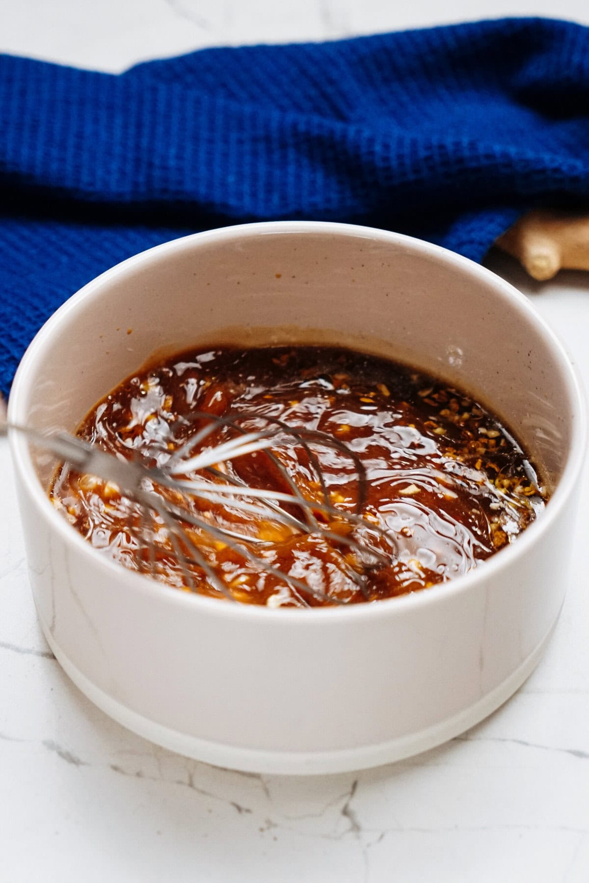 Whisk mixing stir fry sauce in a white bowl on a marble surface, with a blue cloth in the background.