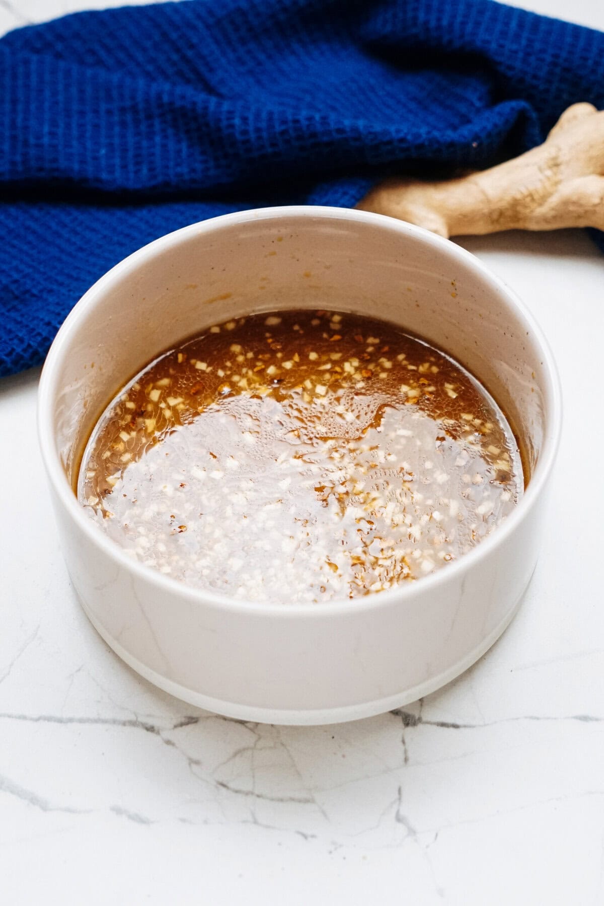 A bowl of rich stir fry sauce featuring minced garlic and sesame seeds rests on a marble surface, with a blue cloth and fresh ginger root nestled in the background.