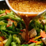 Drizzling stir fry sauce over a vibrant mix of chicken, broccoli, snap peas, carrots, and bell peppers sizzling in the pan.