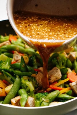 Drizzling stir fry sauce over a vibrant mix of chicken, broccoli, snap peas, carrots, and bell peppers sizzling in the pan.