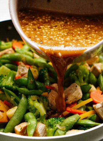 Drizzling stir fry sauce over a vibrant mix of chicken, broccoli, snap peas, carrots, and bell peppers sizzling in the pan.