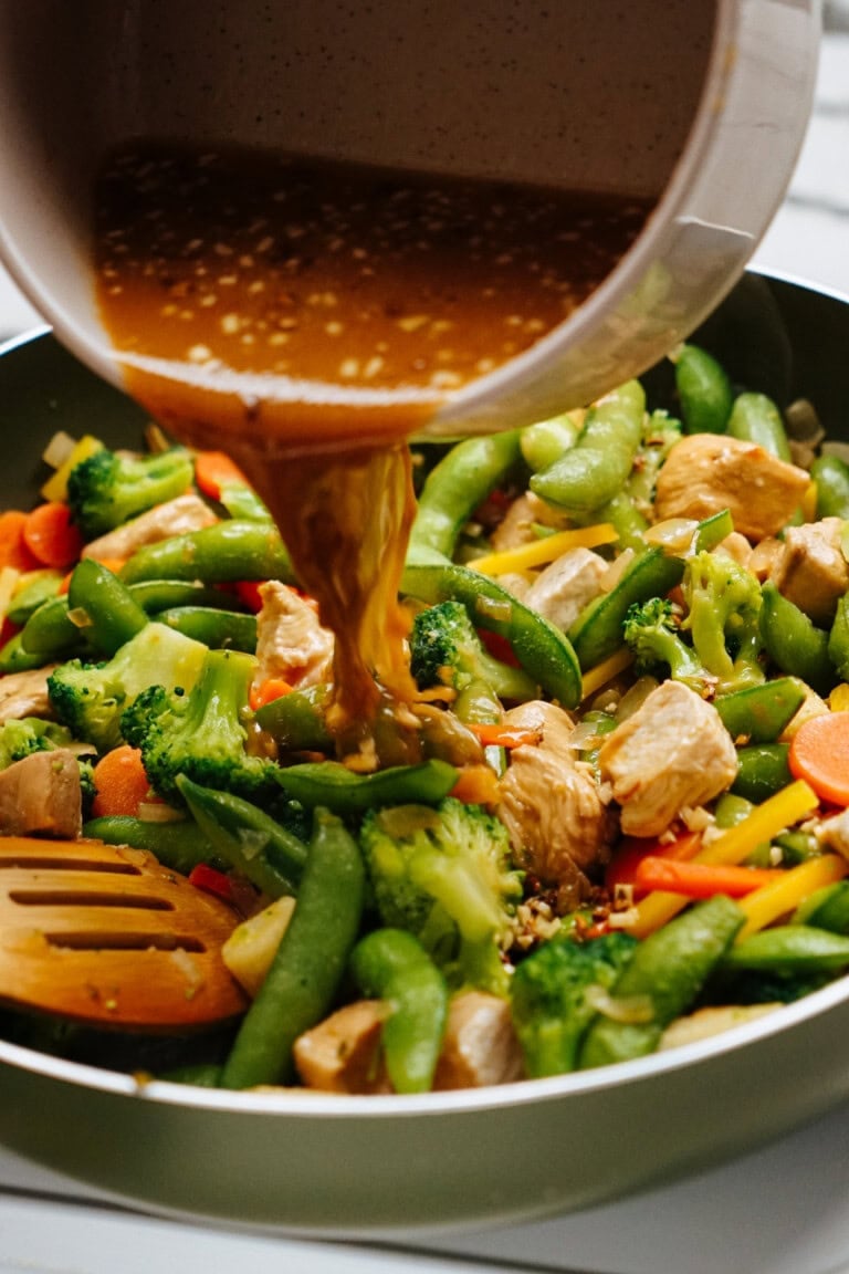 A delicious stir fry sauce cascades into a pan of vibrant vegetables and tender chicken, mingling with broccoli, snap peas, and sliced carrots for a flavorful feast.