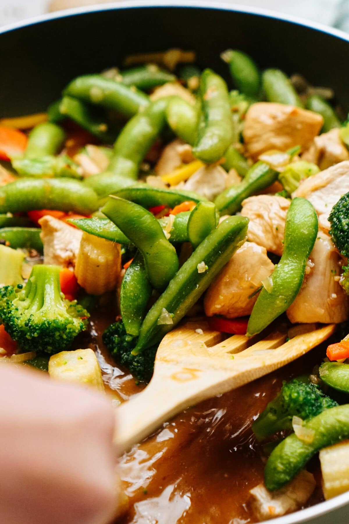 A person stirs a pan of chicken stir-fry with broccoli, snap peas, and red bell peppers using a wooden spatula, ensuring the stir fry sauce is perfectly mixed for a delightful flavor.