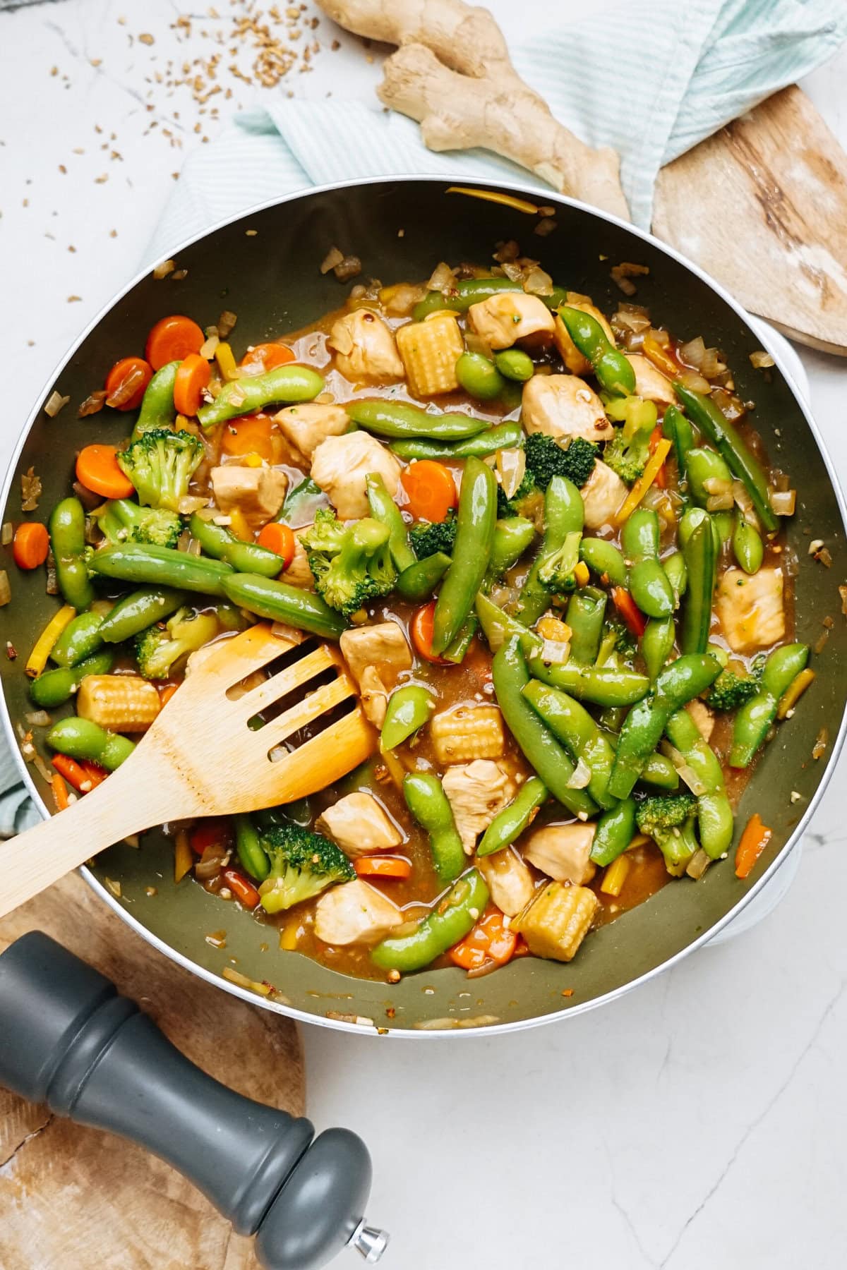 Stir-fried vegetables with chicken and a savory stir fry sauce in a pan, mixed with a wooden spatula on the kitchen counter, where ginger and a cloth are also nearby.