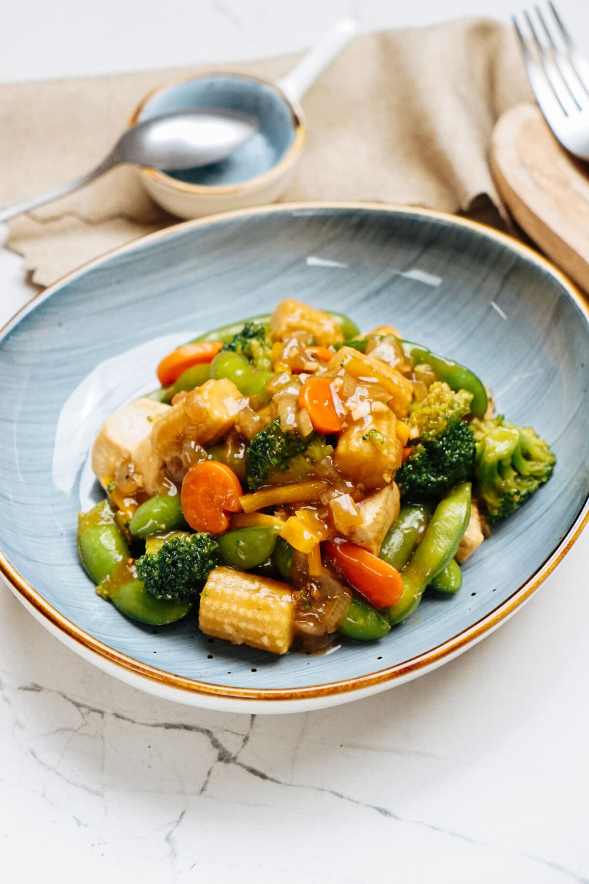 A bowl of stir-fried vegetables with tofu, enhanced by a savory stir fry sauce, includes broccoli, carrots, bell peppers, and baby corn. It's elegantly served on a blue plate with a fork and spoon in the background.