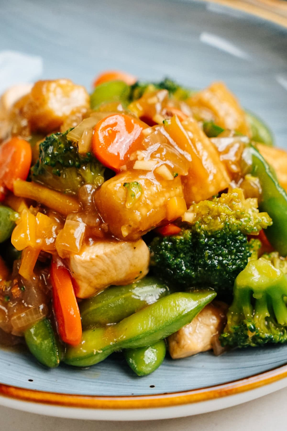 A plate of stir-fried chicken and vegetables, including broccoli, carrots, and snap peas, coated in a glossy stir fry sauce.