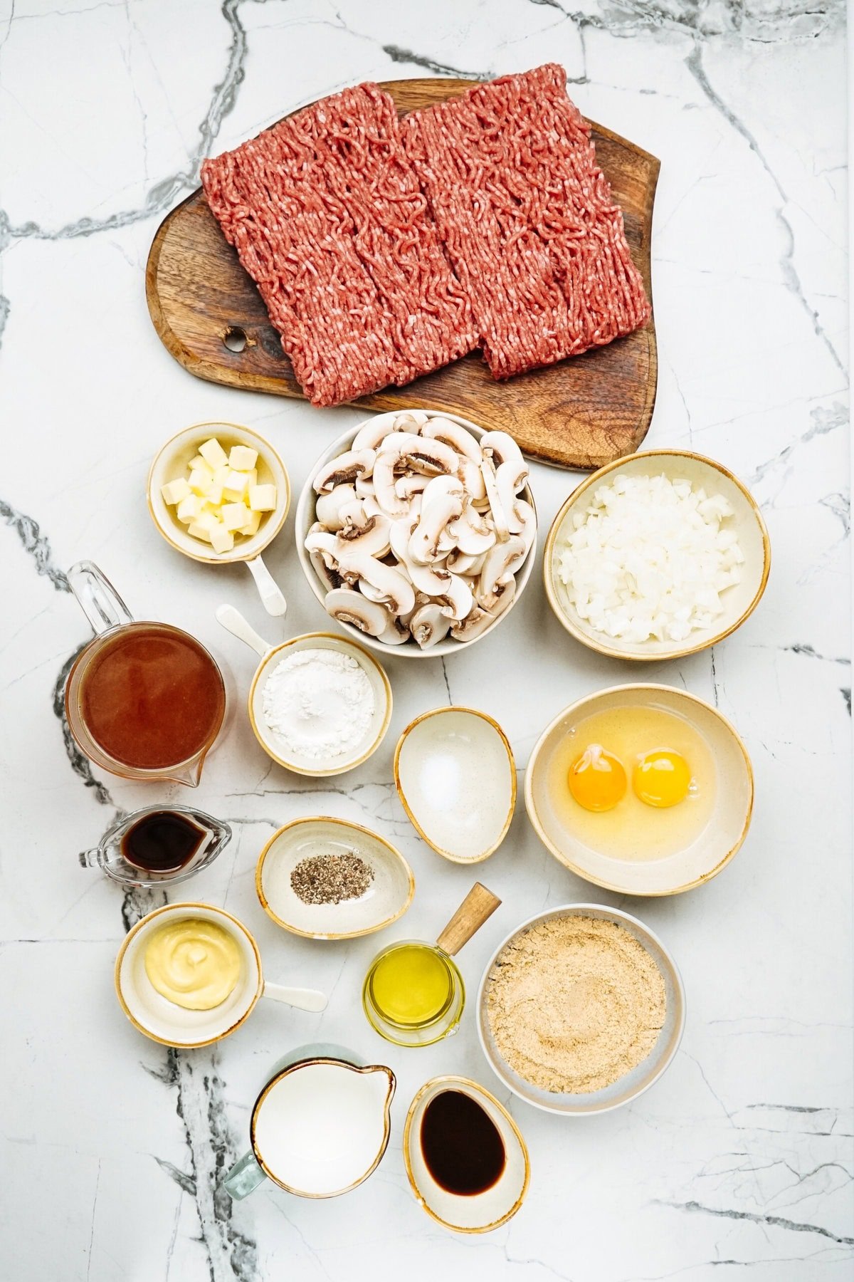 Ingredients for a Salisbury steak recipe on a marble surface: ground beef, sliced mushrooms, diced butter, cooked rice, broth, flour, eggs, sour cream, oil, breadcrumbs, pepper, salt, and a dark liquid.