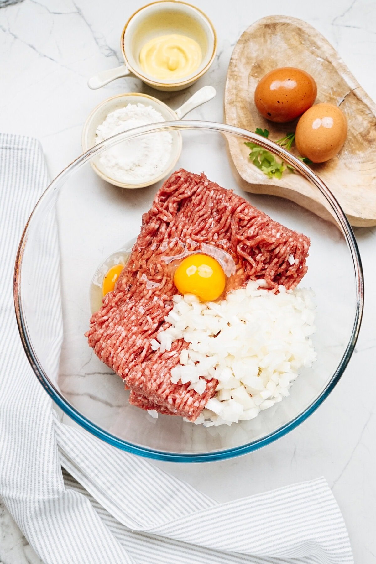 Ground meat, chopped onions, and two raw eggs in a glass bowl are ready for your Salisbury steak recipe. Nearby, flour and mustard sit in small bowls beside two whole eggs on a wooden tray on the marble surface.