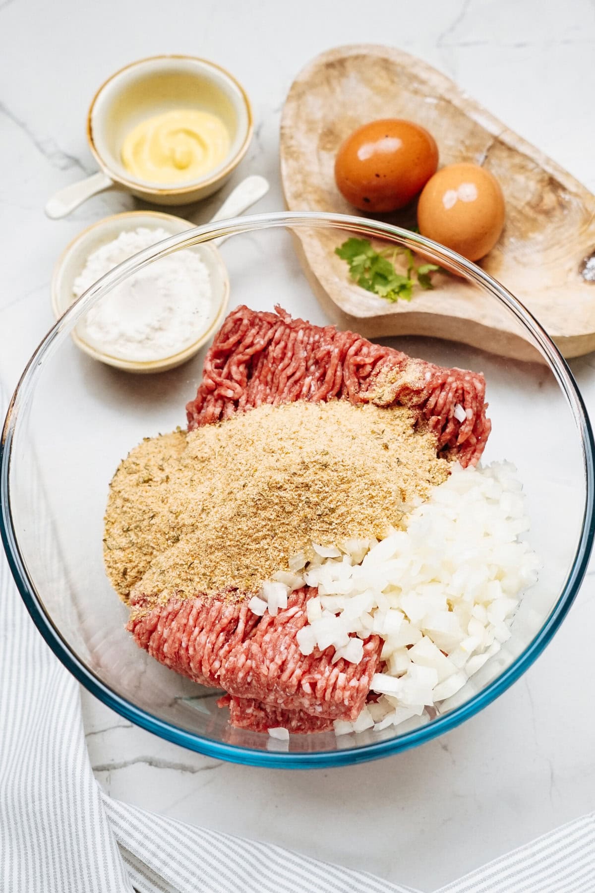 A Salisbury steak recipe awaits creation with ground meat, breadcrumbs, and chopped onions in a bowl. Nearby, eggs, flour, and a saucer of mustard rest on the marble surface.