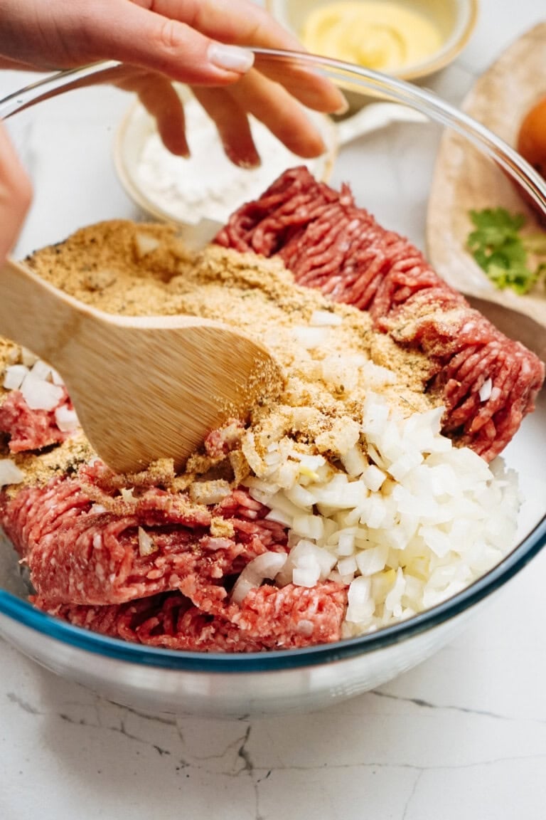 A wooden spoon mixes ground beef, chopped onions, and breadcrumbs in a glass bowl for a delicious Salisbury steak recipe. Other ingredients are visible in the background on a marble counter.