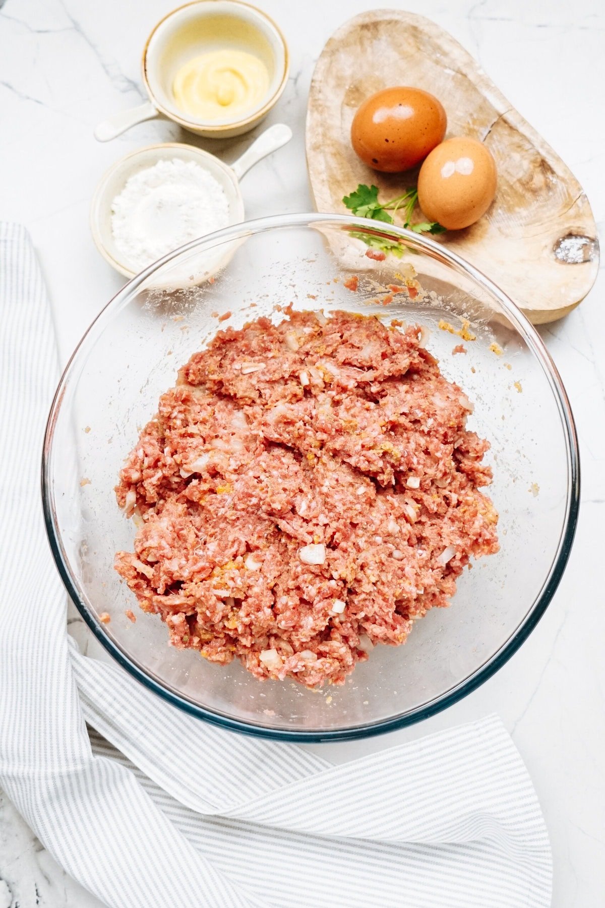 Salisbury steak recipe in progress: ground meat mixture in a glass bowl on a countertop, surrounded by eggs on a wooden tray, small bowls of butter and flour, and a striped kitchen towel nearby.