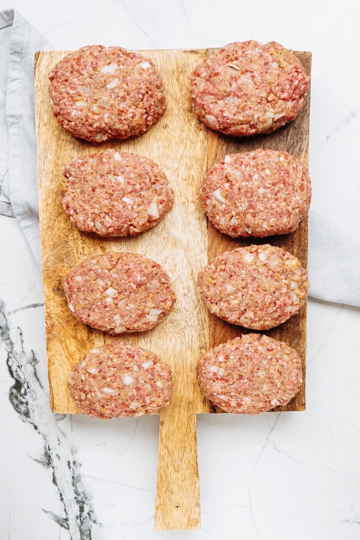 Eight raw hamburger patties, perfect for a savory Salisbury steak recipe, are arranged on a wooden cutting board placed on a marble countertop.