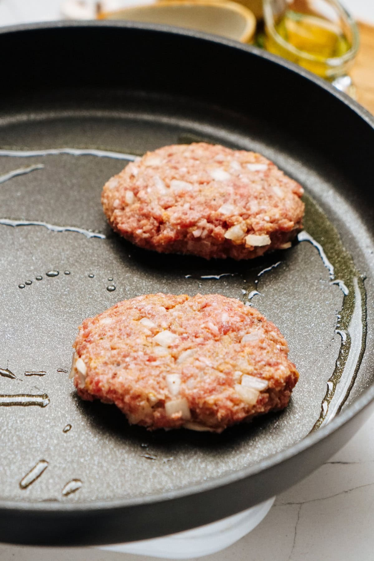 Two raw burger patties with onions are cooking in a black frying pan with a touch of oil, reminiscent of a classic Salisbury steak recipe.