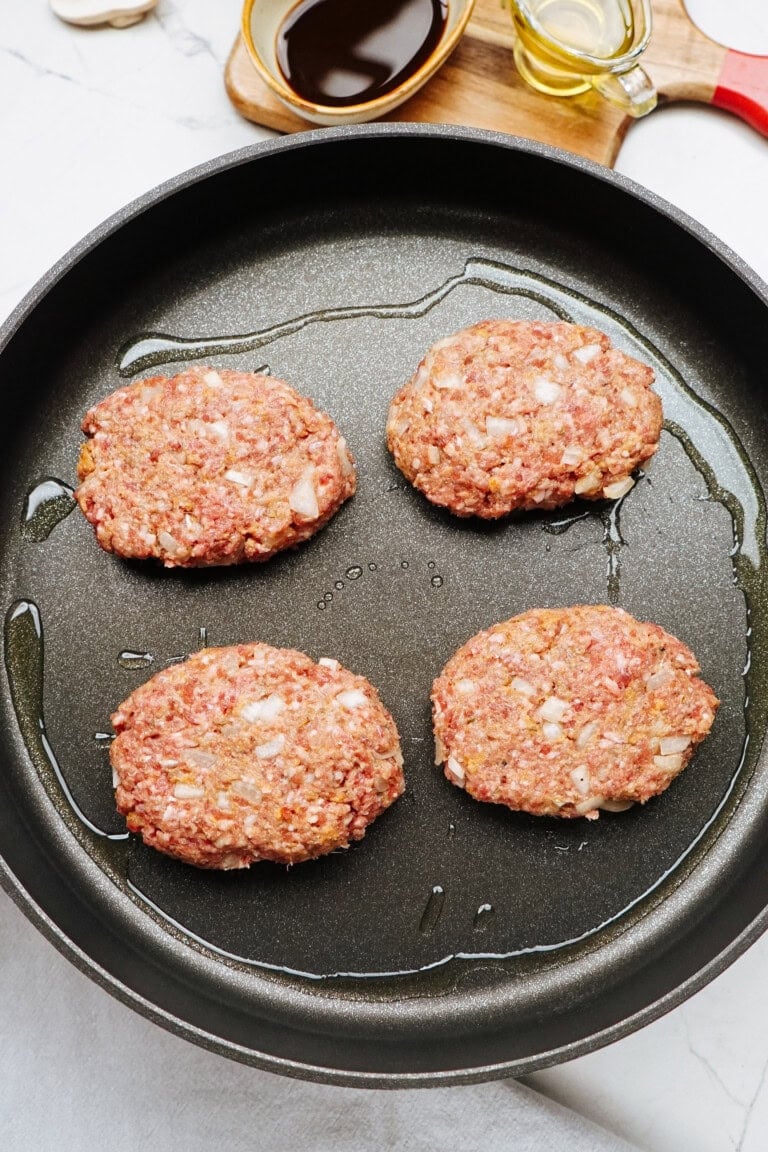 Four raw burger patties with diced onions sizzle in a frying pan, surrounded by oil. In the background, a small container of oil and soy sauce hints at a savory Salisbury steak recipe waiting to be created.