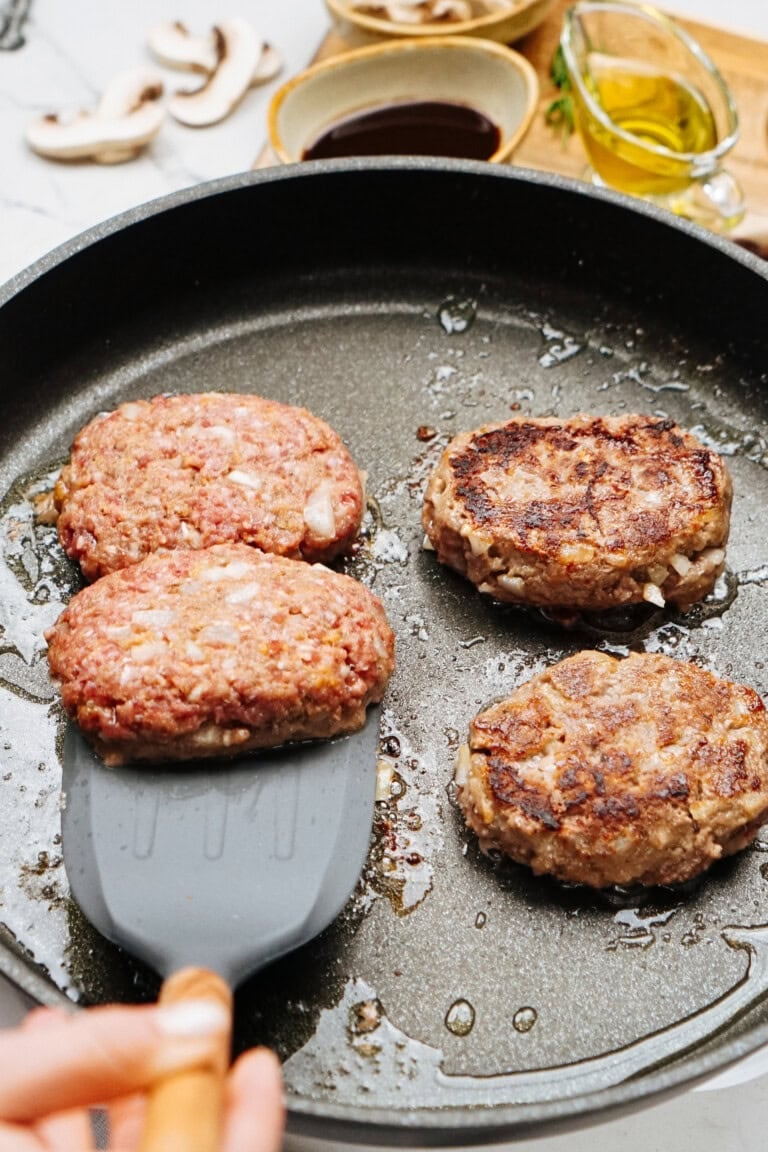 Four beef patties sizzle in a frying pan, reminiscent of a classic Salisbury steak recipe, as a spatula gracefully lifts one. In the background, ingredients neatly arranged in small bowls hint at a delicious creation underway.