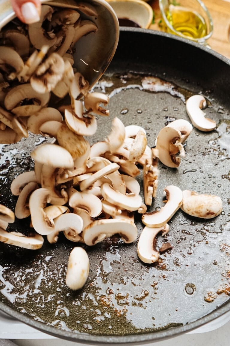 Sliced mushrooms are being added to the frying pan on the stovetop, enhancing the savory goodness of your Salisbury steak recipe.