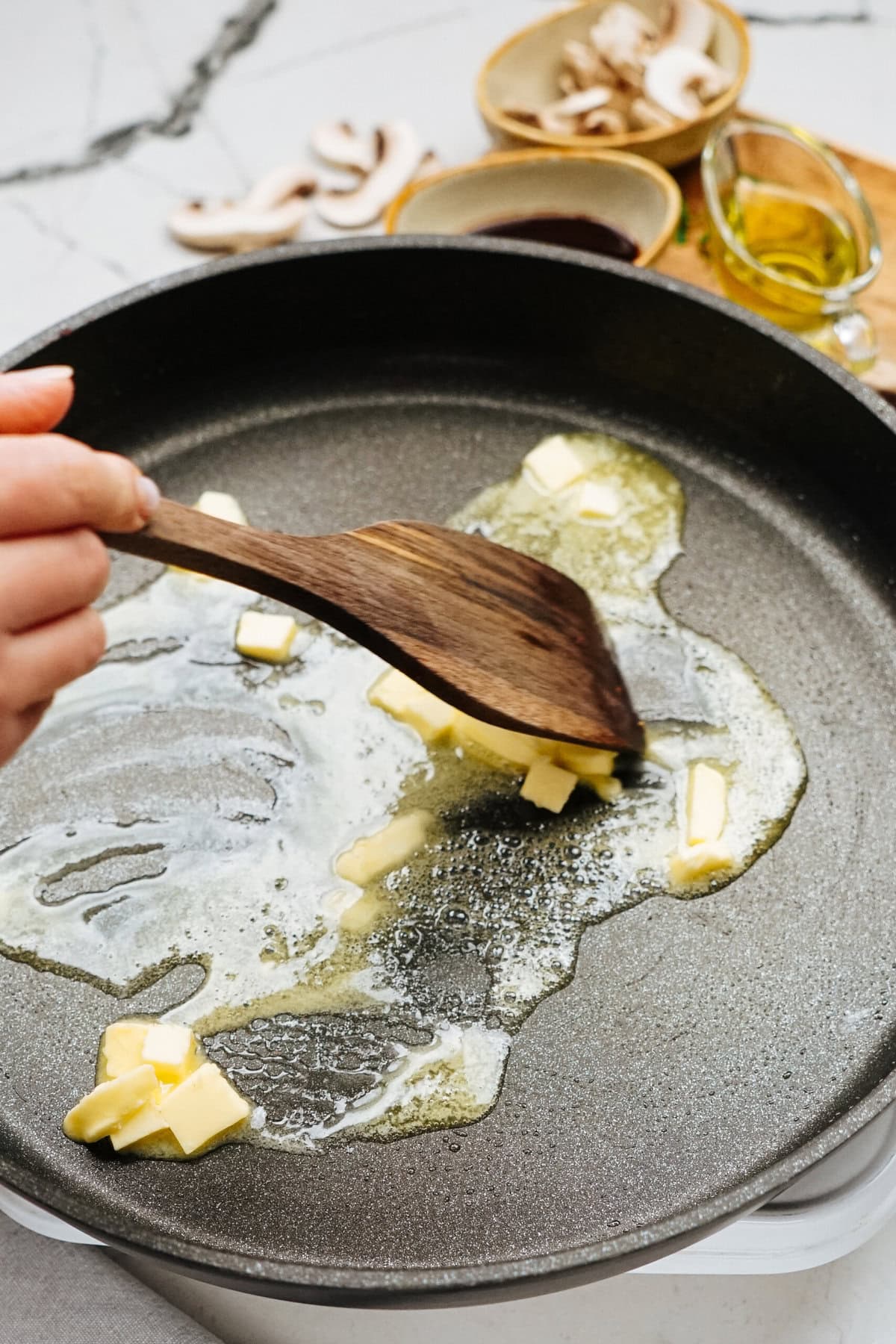 Close-up of a hand using a wooden spatula to stir melting butter in a frying pan, perfectly setting the stage for our Salisbury steak recipe. Bowls of mushrooms, sauce, and olive oil sit ready in the background, promising rich flavor and hearty satisfaction.