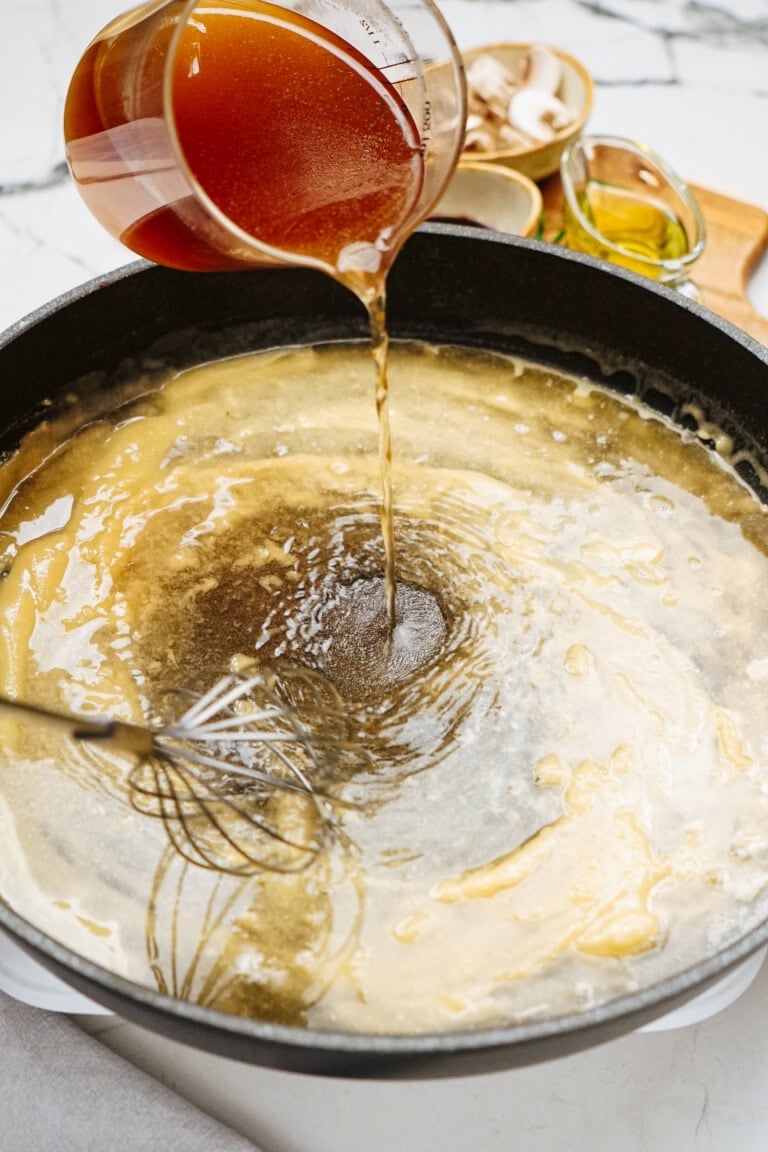 Broth being poured into a pot with a whisk inside, surrounded by various ingredients on a white countertop, sets the stage for crafting the perfect Salisbury steak recipe.