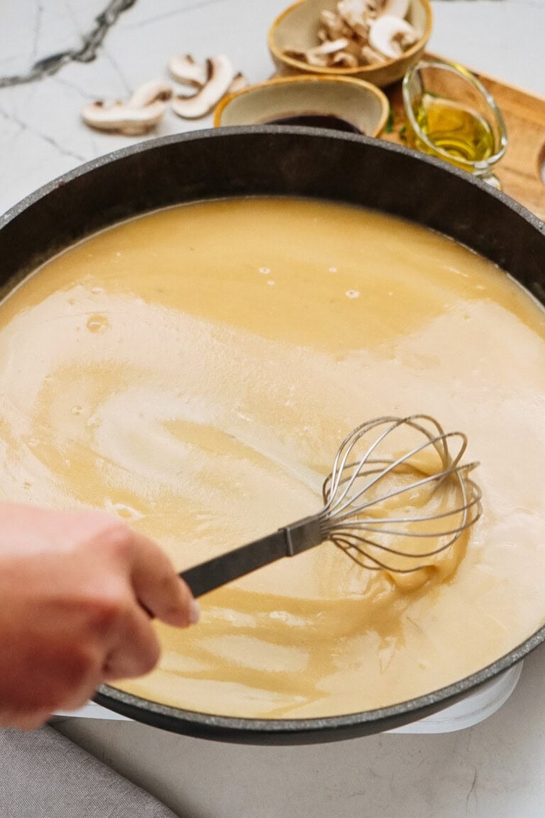 Hand whisking a creamy sauce for a classic Salisbury steak recipe in a large black pan, with sliced mushrooms and oil in small bowls nearby on a marbled surface.