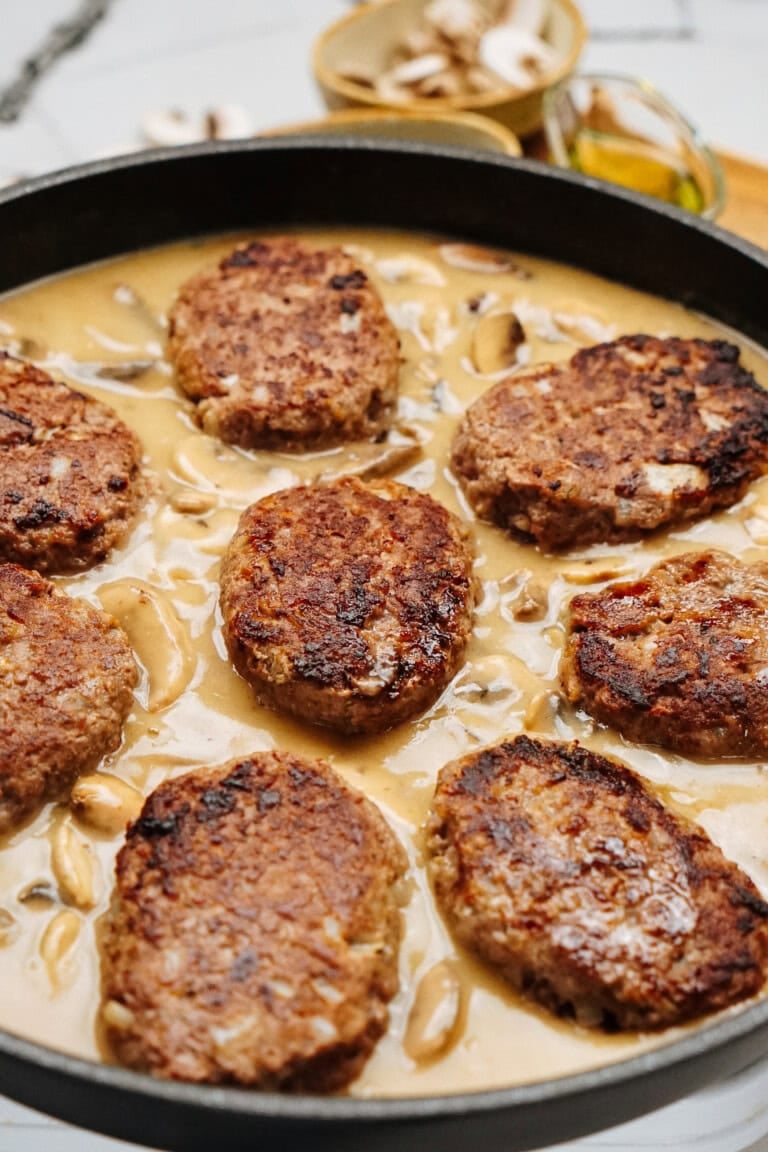 A pan filled with seven browned meat patties simmering in a creamy mushroom sauce creates a classic Salisbury steak recipe, garnished with freshly sliced mushrooms.