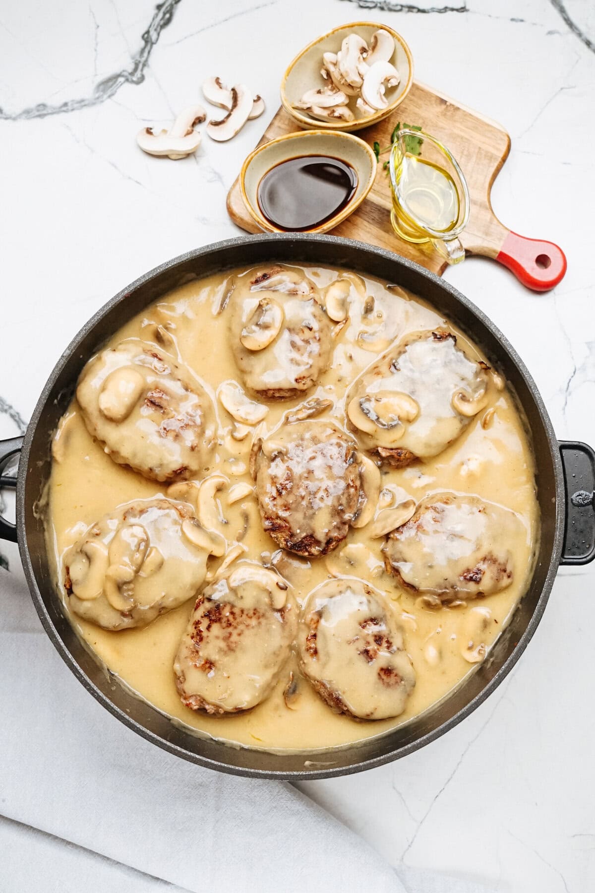 A skillet with cooked hamburger steaks in a creamy mushroom sauce, reminiscent of a classic Salisbury steak recipe, is garnished with mushroom slices. Bowls of fresh mushrooms, olive oil, and soy sauce rest on a wooden board nearby.