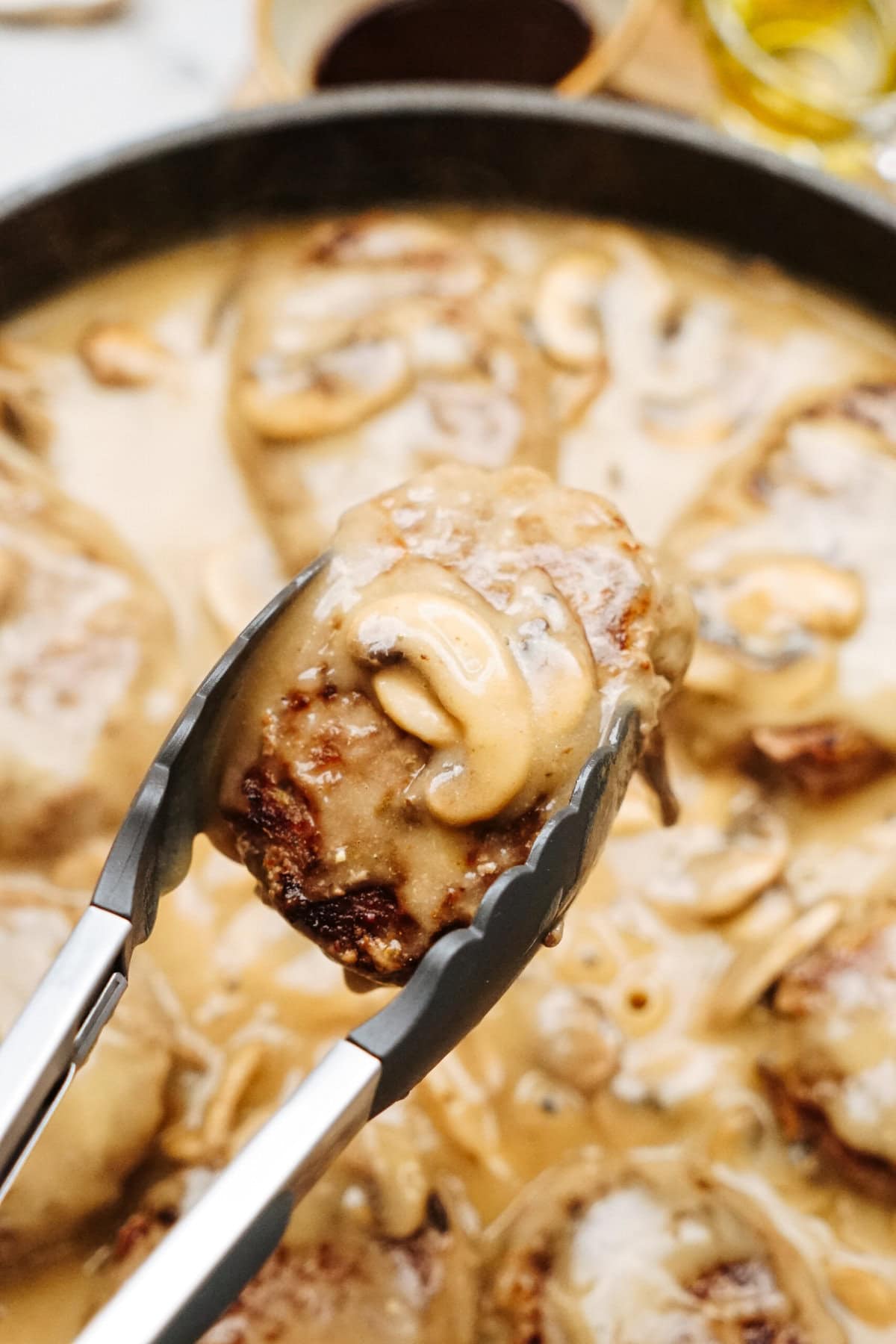 Tongs holding a piece of meat covered in mushroom sauce over a pan filled with more pieces in savory Salisbury steak recipe sauce.