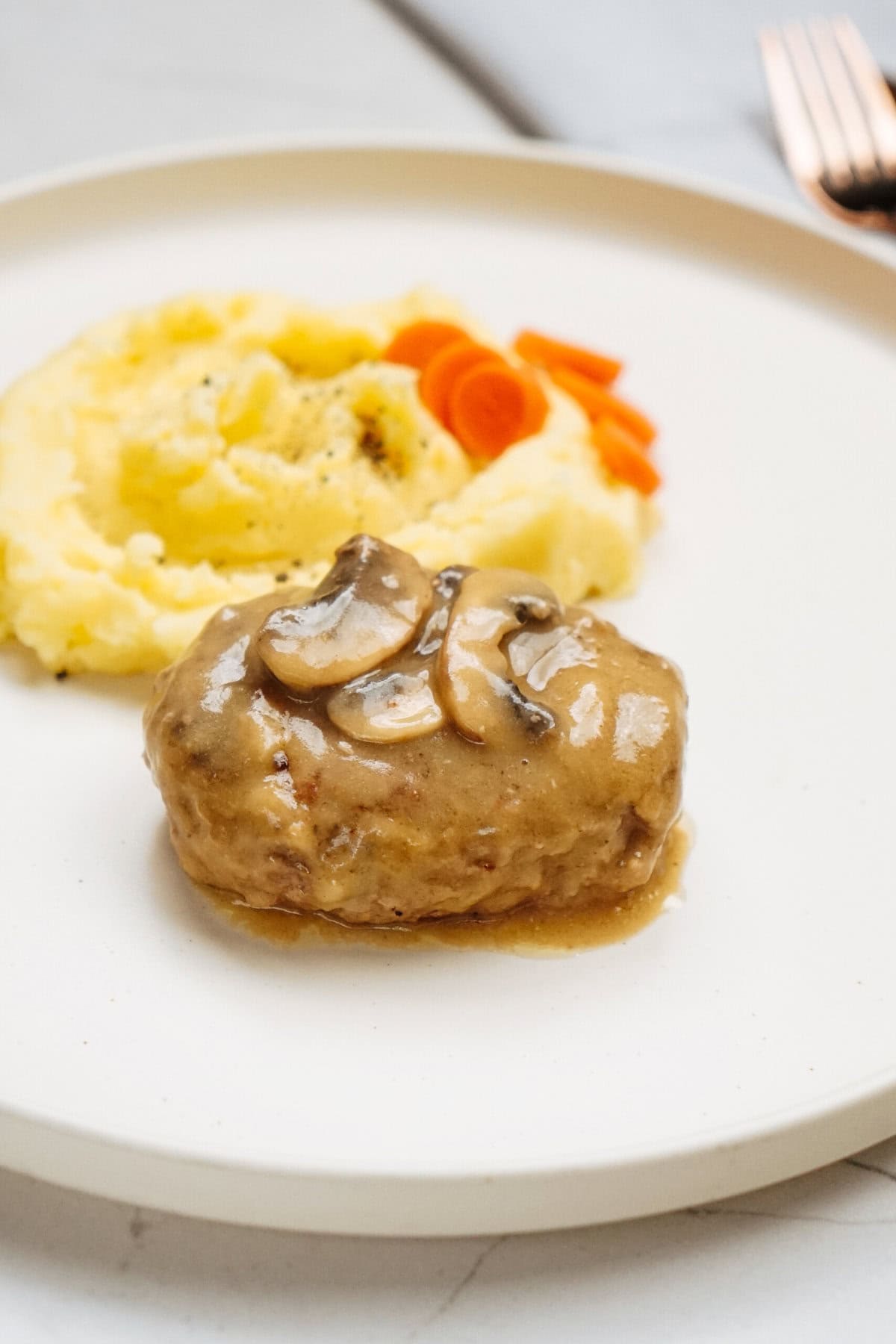 A plate featuring mashed potatoes, sliced carrots, and a tender piece of meat smothered in mushroom gravy offers a delightful taste of a classic Salisbury steak recipe.