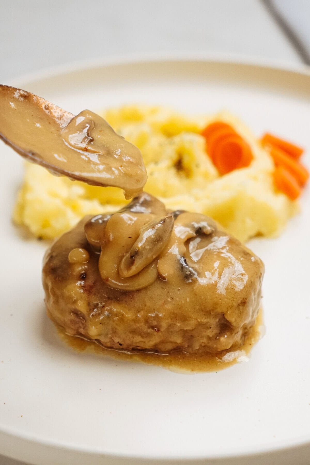 Gravy with mushrooms being poured over a serving of meatloaf on a white plate resembles a classic Salisbury steak recipe, complemented by mashed potatoes and sliced carrots.