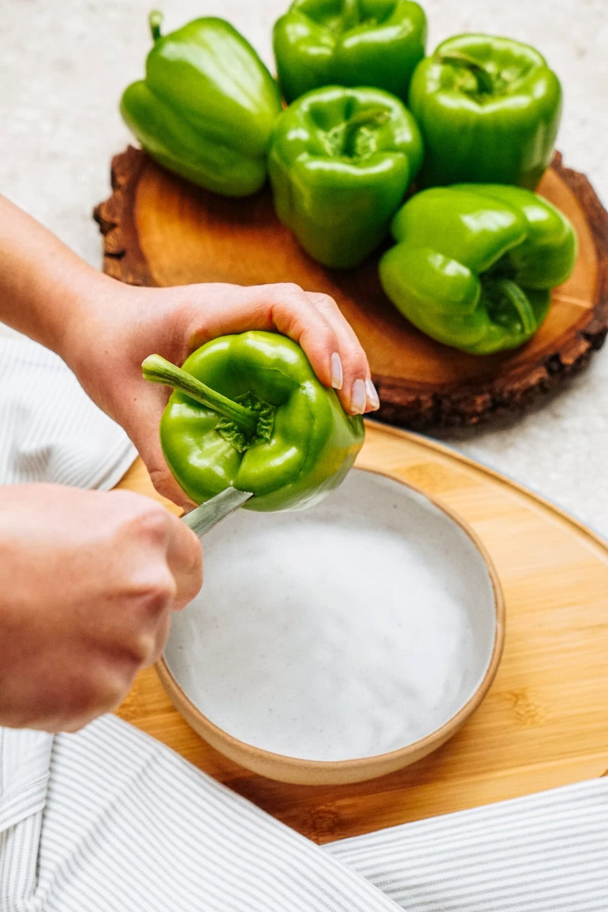 A person carefully cuts a green bell pepper in half above a bowl, preparing for stuffed peppers. More vibrant bell peppers rest on a wooden board in the background.