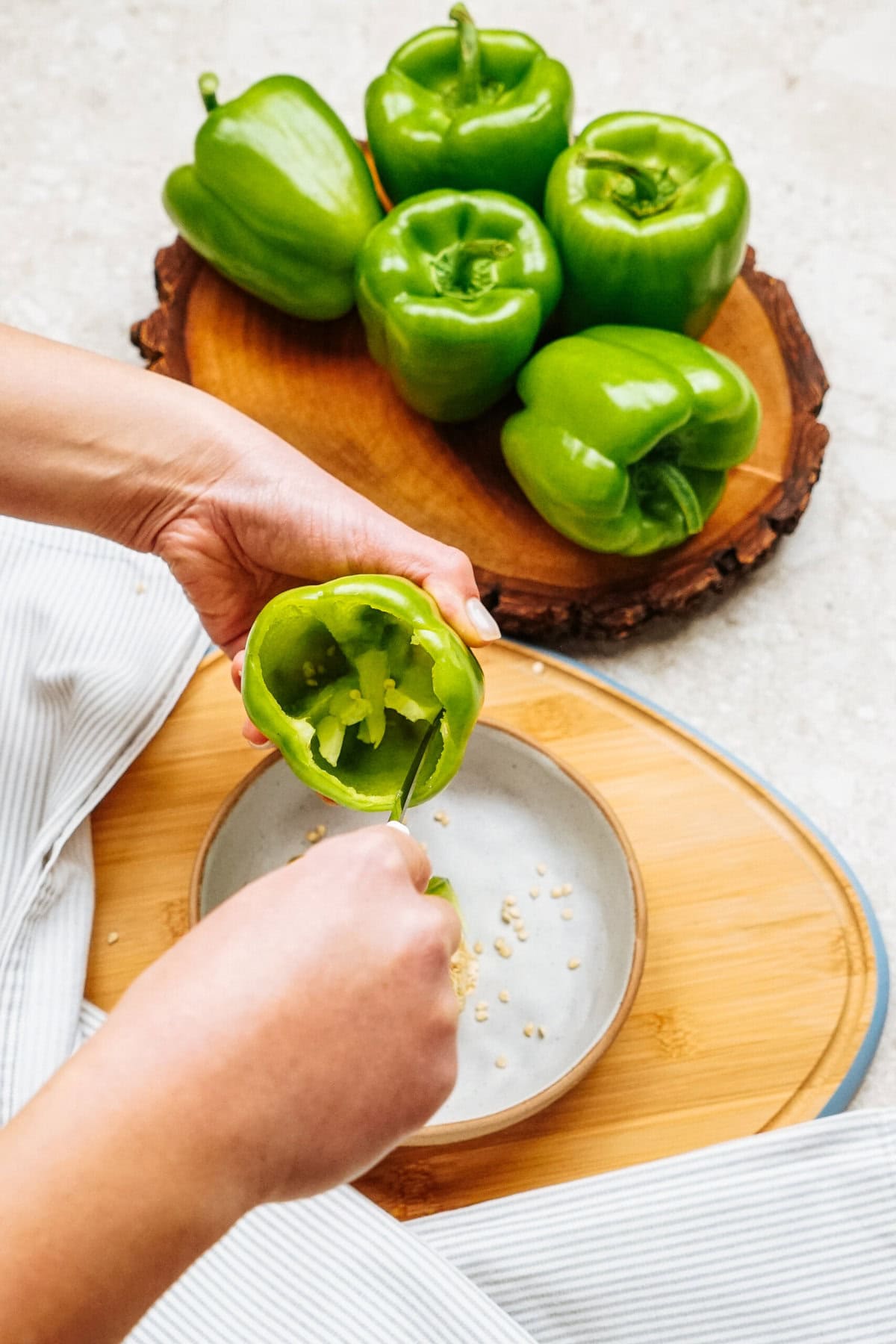 A person is using a spoon to remove seeds from a green bell pepper, prepping it for delicious stuffed peppers. Several whole green bell peppers sit on a wooden tray, ready for the next step.