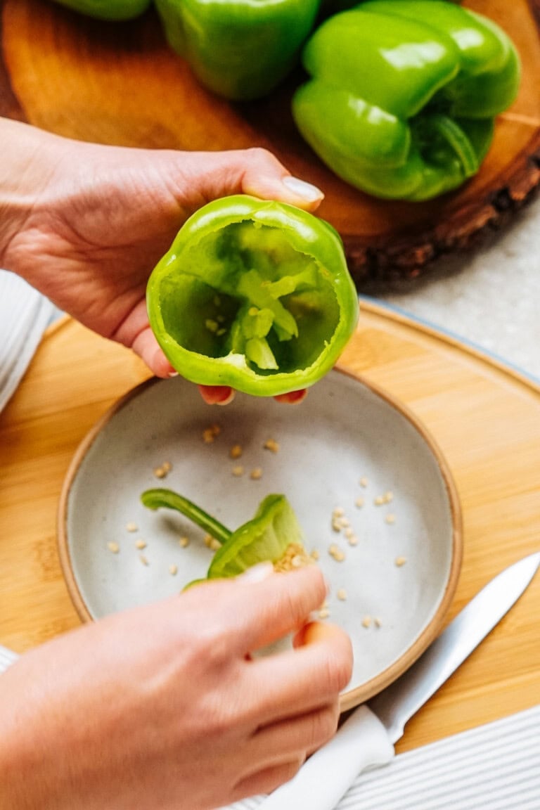 Hands are holding a green bell pepper above a bowl, preparing it for stuffed peppers by removing seeds with a knife placed nearby. Other green bell peppers await in the background on a wooden surface.