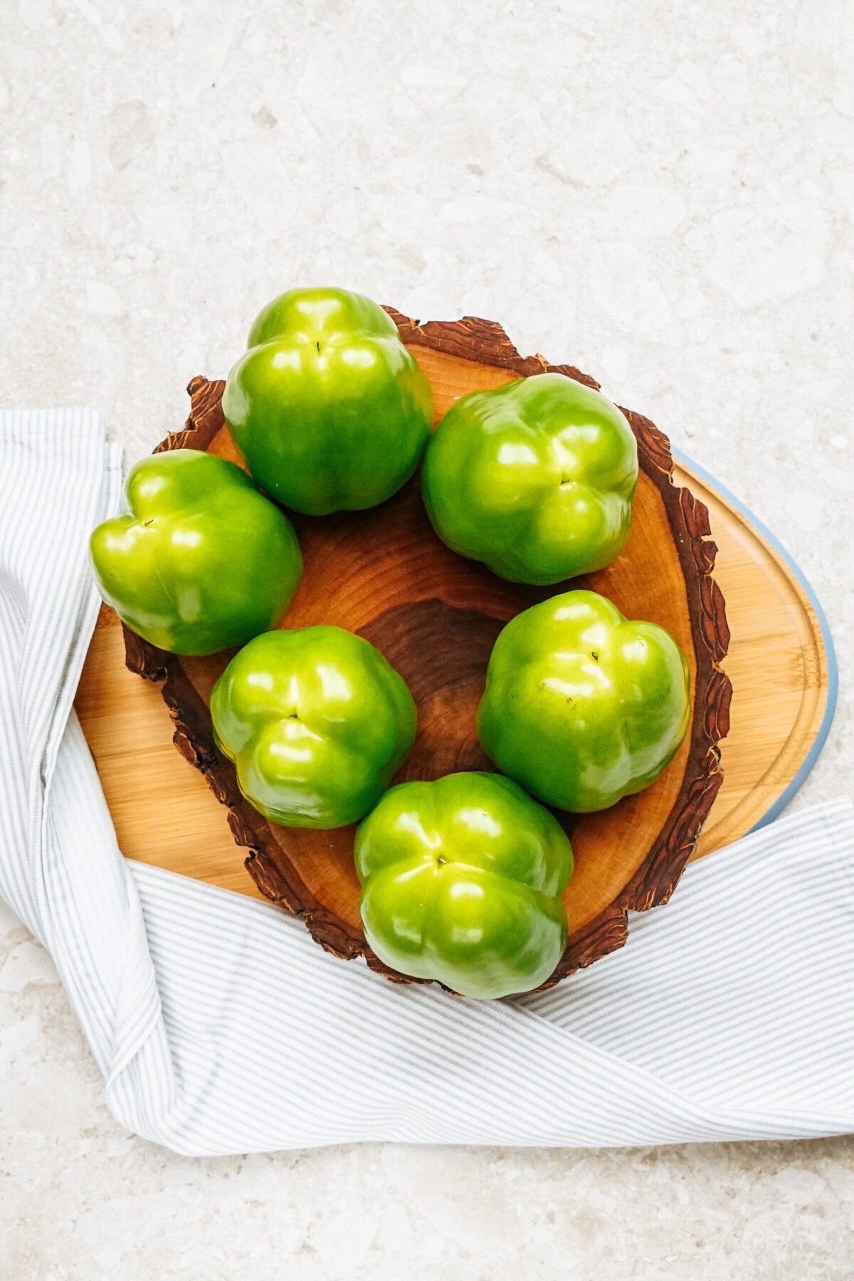 Six vibrant green bell peppers are arranged in a circle on a wooden platter, hinting at their potential as delicious stuffed peppers, all set against the backdrop of a striped cloth on a textured surface.