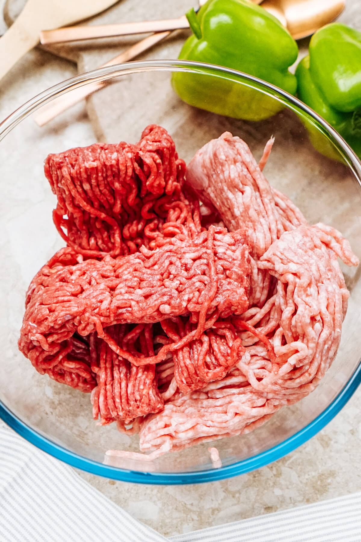 Ground meat in a glass bowl on a countertop, with green bell peppers in the background, perfect for preparing stuffed peppers.