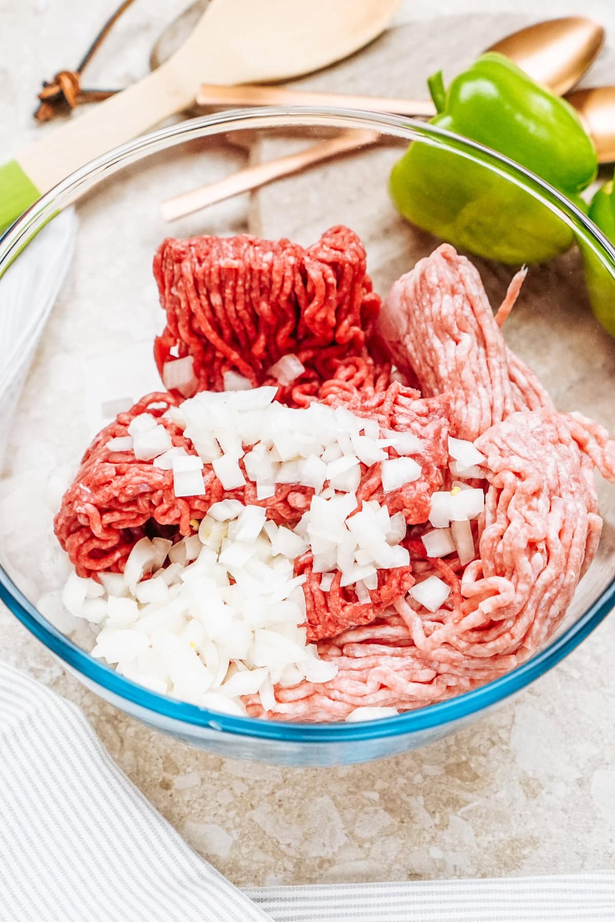 A glass bowl containing raw ground beef mixed with chopped onions rests on a light-colored surface, ready for creating delicious stuffed peppers. Nearby, a green bell pepper and wooden utensils await their turn in this culinary adventure.