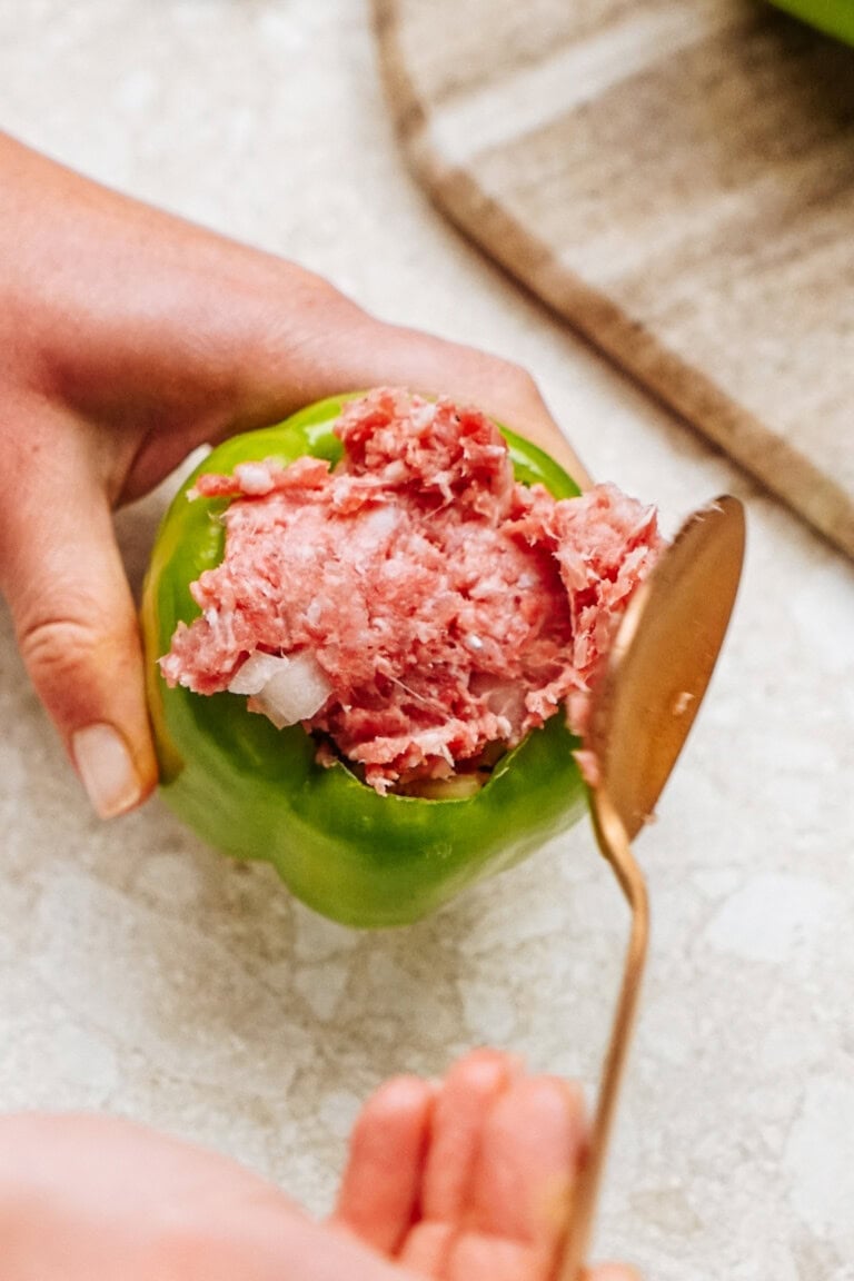 A hand expertly fills a green bell pepper with a savory blend of ground meat and onions, crafting perfect stuffed peppers using a spoon.