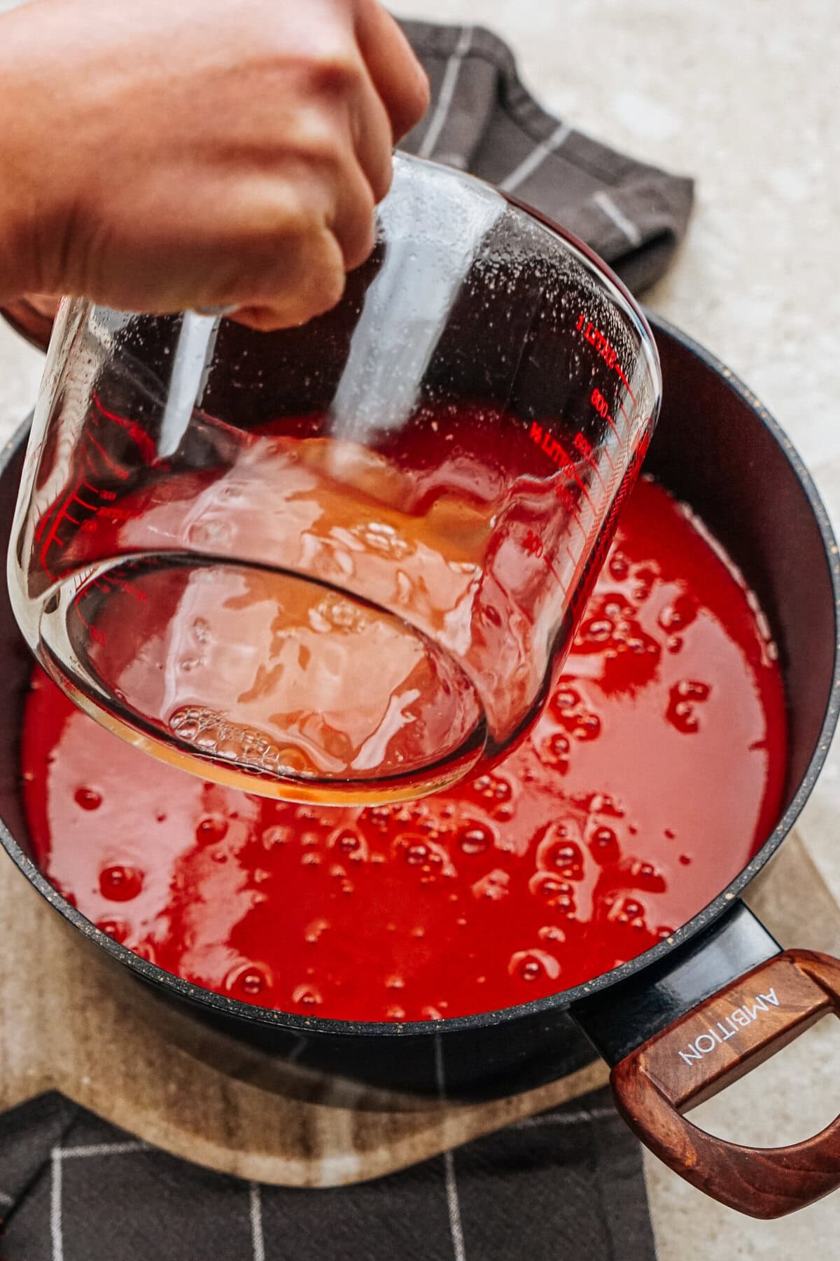 Hand pouring liquid from a measuring cup into a pot of tomato sauce on the stove, creating the perfect base for flavorful stuffed peppers.