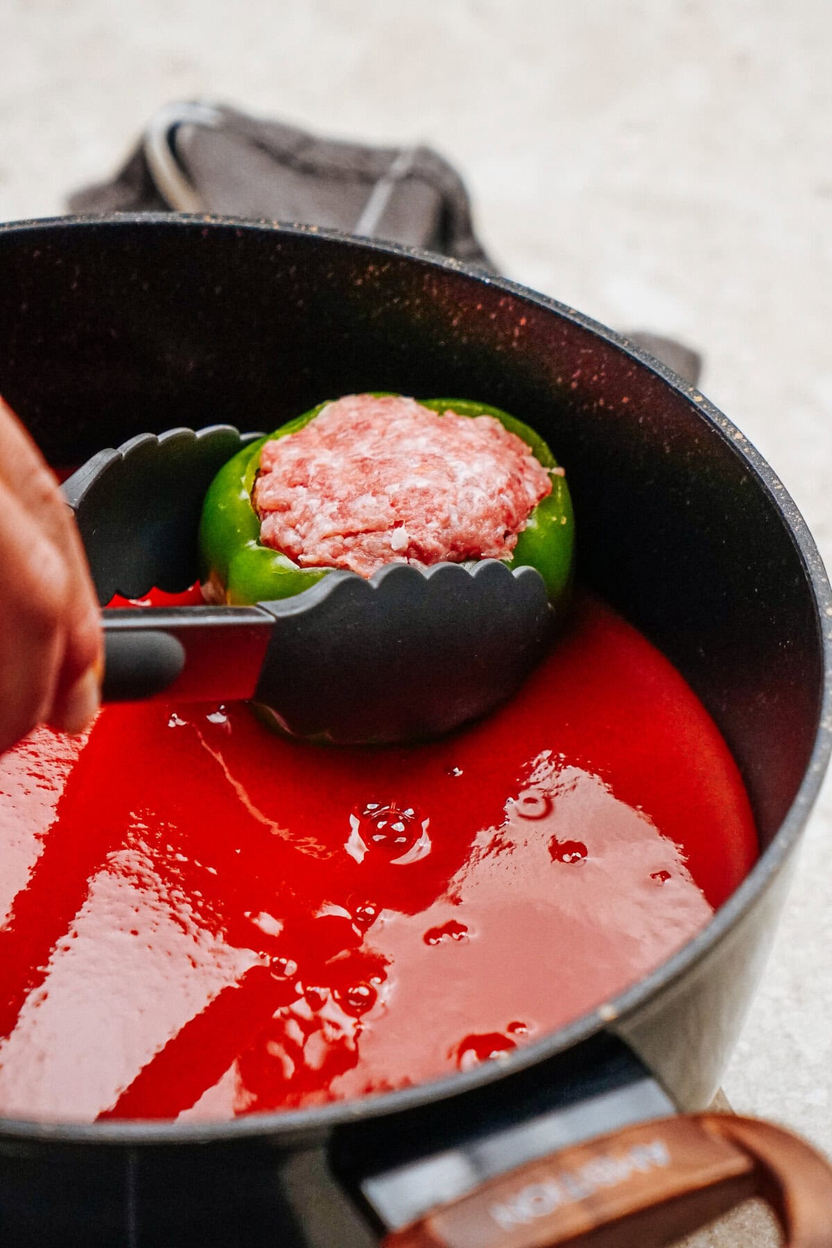 Tongs holding a green stuffed pepper filled with ground meat hover over a pot brimming with rich red sauce.