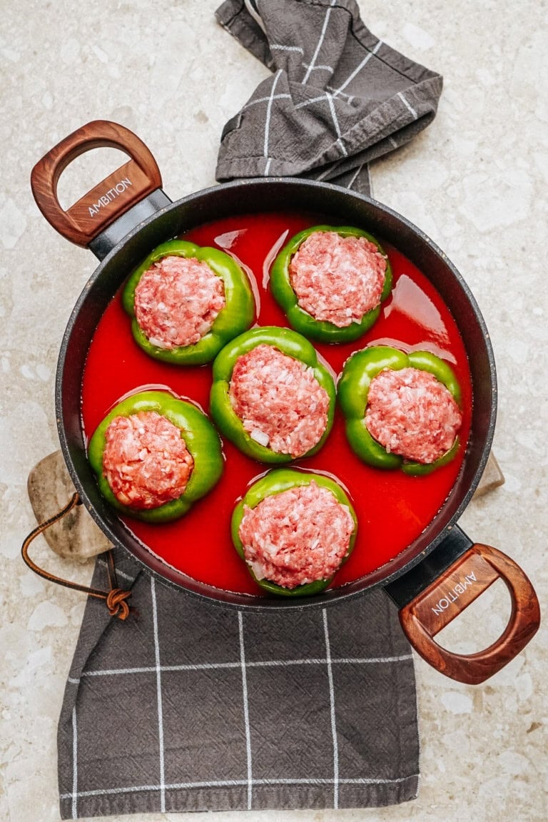 Stuffed peppers with ground meat rest in a red sauce-filled pan, elegantly placed on a gray checkered cloth.