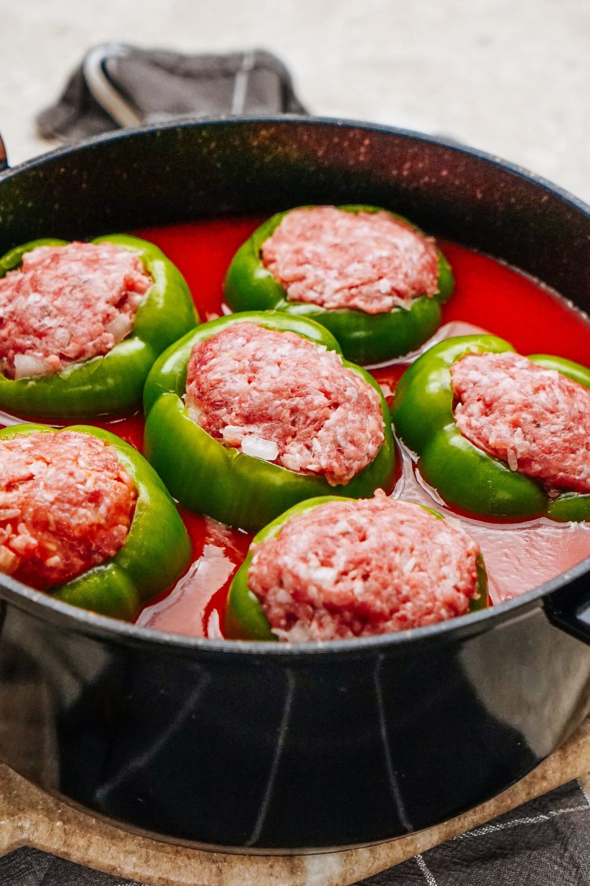 Stuffed peppers with raw ground meat are arranged in a black pot filled with rich tomato sauce.