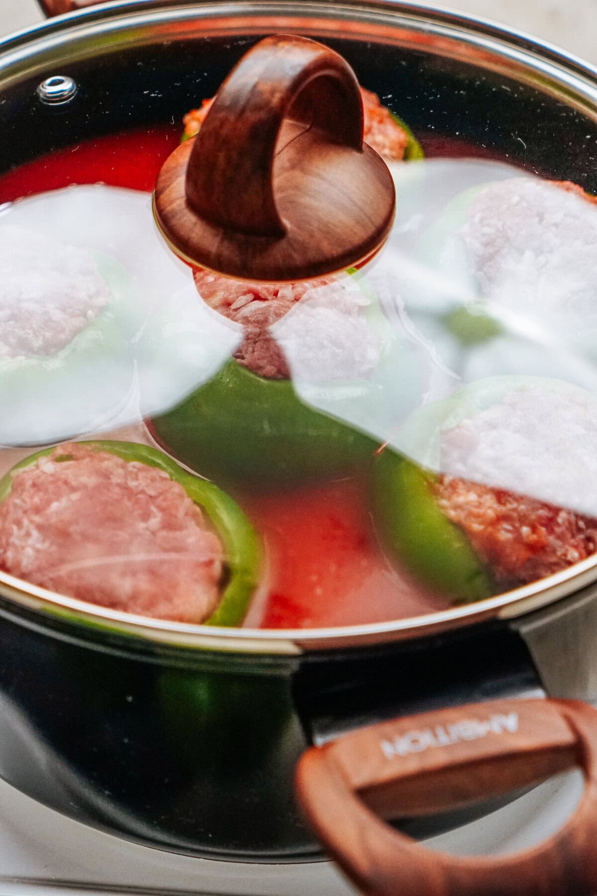 A pot with a wooden handle lid sits on the stovetop, brimming with stuffed peppers. Green bell peppers are filled with savory meat, simmering gently in a rich red liquid.