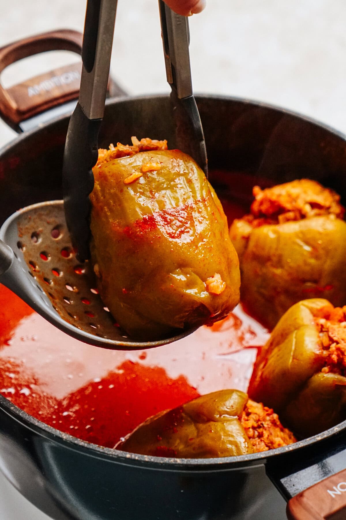 A stuffed pepper is being lifted from a pot with red sauce using tongs. .